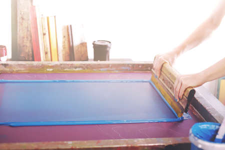 a big silkcreen and a aluminum squeegee are used to print a graphic on fabric with blue paint by a beautiful woman with matching painted nails - bright light mood and selective focusの写真素材
