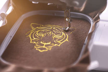 modern embroidery machine stitches a luxurious tiger head on brown merino wool fabric with golden...