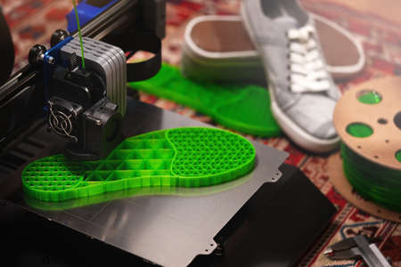 vivid green shoe sole is printed from translucent filament on a cartesian 3D printer with steel print bed. comfy home surrounding with persian rug, gray sneaker shoes and filament. selective focusの写真素材
