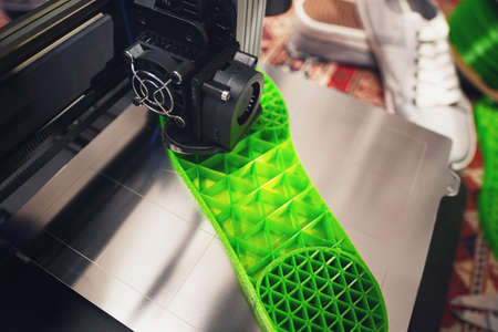 3D printer produces a shoe sole with distinct inner structure from green plastic filament. top view closeup with steel print bed, gray sneaker shoes, filament spool and persian rug. selective focusの写真素材