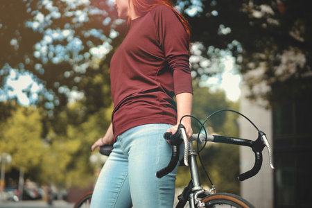 red-haired woman in tight blue jeans stands in front of a vintage bicycle holding handlebar and saddle. selective focus. part view on front of upper body, hips and thighs. urban lifestyle conceptの写真素材