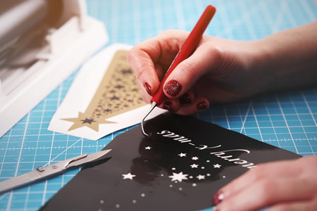 close up on weeding process of christmas graphic cut from black vinyl foil. weeding tool, scalpel and fermale hands with red nails. background blanked out blurry. gift and advertising conceptの写真素材