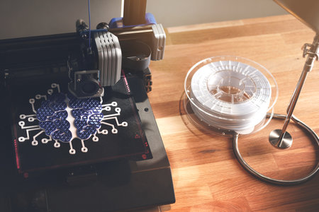 FDM 3D printer on wooden working surface prints a brain sculpture from blue and white plastic. Tidy work space with desk lamp and filament roll in bright light. selective focus on print headの写真素材
