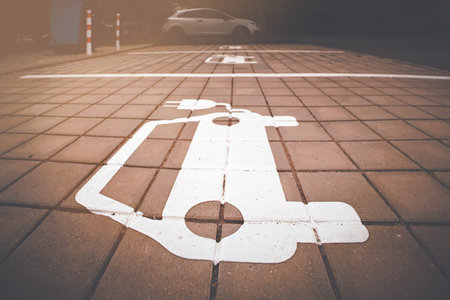 Electric vehicle parking symbol on paved charging space with warm sunlight and parked car in the background.の写真素材