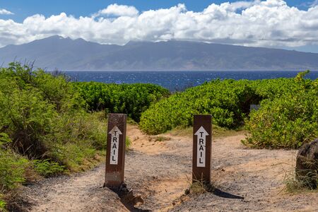 Trail Signs With White Straight Ahead Arrows on Posts Pointing Towards Ocean Water and Island in the Distanceの写真素材