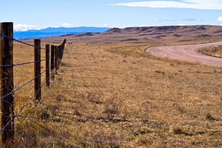 A barbed wire fence runs along a gravel road, east of the Rocky Mountainsの写真素材