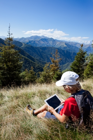 Young boy sitting in a mountain meadow using a tablet computer. Summer season, clear blue sky. Boy is wearing a red shirt, white cap and a grey backpack. Piemonte, West Italian Alps.の写真素材