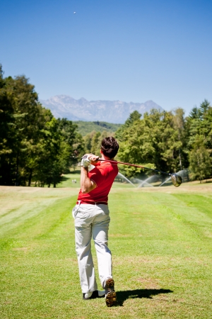 Golf player performs a tee shot using a driver club. One young caucasian male golfer, red shirt and white pants. Clear summer day, rear view. Summer outdoor sport and wealthy class concept.の写真素材