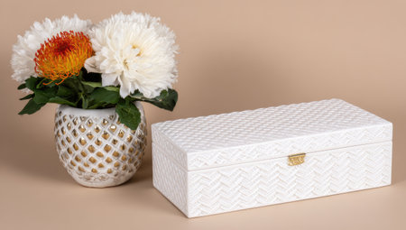 White decorative box with a woven pattern. A gold-detailed, ceramic, patterned vase holding a bouquet of white and orange flowers sits beside the box. The items are against a neutral beige background.の素材