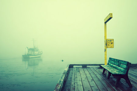 Tranquil harbor scene on a foggy day. A small boat floats on the still water of the harbor, and a weathered wooden bench sits on a wooden pier stretching into the misty expanse.の素材