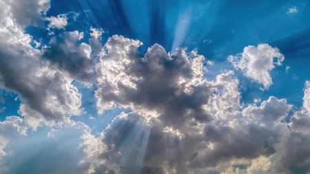 Dramatic, wide-angle view looking up at a vibrant blue sky densely populated with large, contrasting clouds, where intense shafts of sunlight emanate from behind the dense formations, creating noticeable crepuscular rays slicing through the atmosphere.の素材
