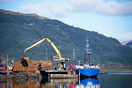 Crane lifting wood logs from stack to ship in sea loch for transportation of biomass fuel in...