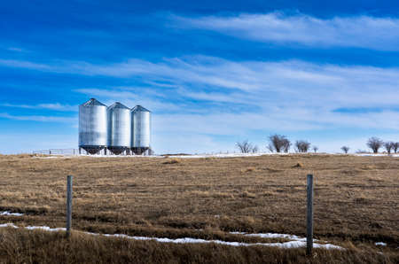 Grain silos holding wheat standing on natural grass lands surrounded by a barbed wire fence on the Canadian prairies in Rocky View County Alberta under a morning sky.の写真素材
