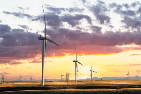 Windmills at sunset producing green energy overlooking agriculture wheat fields on a prairie landscape with distant mountains under a dramatic colourful sky.の写真素材