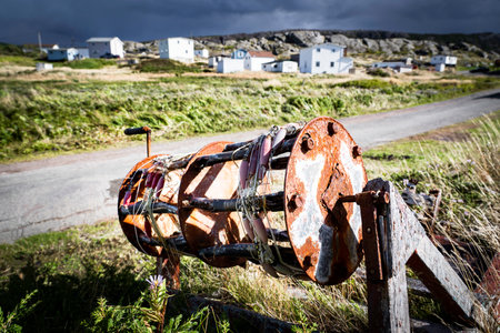 Rustic hand reel from a commercial fishing boat lying in tall grass overlooking small wooden homes at Keels Newfoundland Canada along the Discovery Trail scenic route.の写真素材