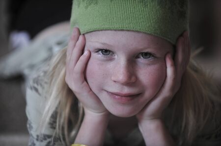 Little girl with her head in her hands, wearing a green wool hat, gazing into the camera.の写真素材