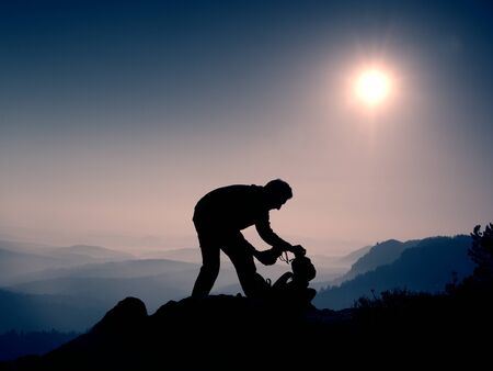 Professional photographer is packing camera into backpack on peak of rock. Dreamy fogy landscape, spring orange pink misty sunrise in a beautiful valley belowの写真素材