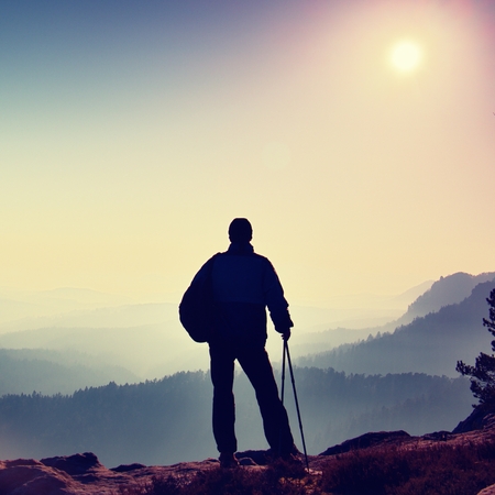 Silhouette of tourist with poles in hand. Hiker with sporty backpack stand on rocky view point above misty valley. Sunny spring daybreak in rocky mountains.の写真素材
