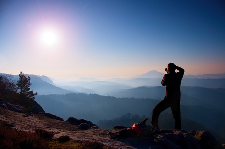 Professional photographer takes photos with mirror camera on peak of rock. Dreamy fogy landscape, spring orange pink misty sunrise in a beautiful valley belowの写真素材