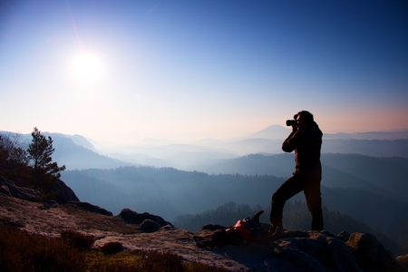 Professional photographer takes photos with mirror camera on peak of rock. Dreamy fogy landscape, spring orange pink misty sunrise in a beautiful valley belowの写真素材
