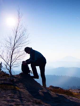 Professional photographer is packing camera into backpack on peak of rock. Dreamy fogy landscape, spring orange pink misty sunrise in a beautiful valley belowの写真素材