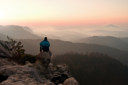 Tourist in blue wind jacket sit on rocky view point and watching into deep misty valley bellow. Sunny spring daybreak in rocky mountains.の写真素材