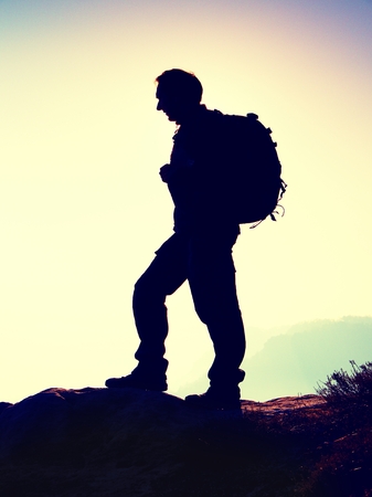 Tourist with sporty backpack stand on rocky view point and watching into deep misty valley bellow. Sunny spring daybreak in rocky mountains.の写真素材