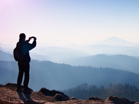 Tourist takes photos with smart phone on peak of rock. Dreamy fogy landscape, spring orange pink misty sunrise in a beautiful valley belowの写真素材