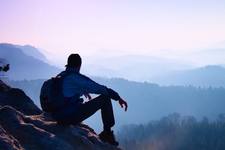 Tourist with sporty backpack sit on rocky peak  and watching into deep misty valley bellow. Sunny spring daybreak in rocky mountains.の写真素材