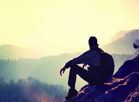 Tourist with sporty backpack sit on rocky peak  and watching into deep misty valley bellow. Sunny spring daybreak in rocky mountains.の写真素材