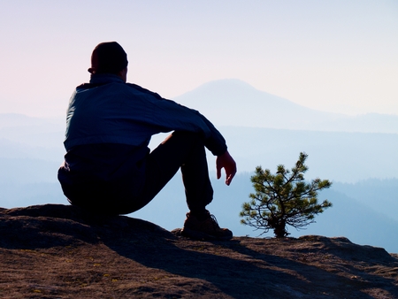 Tourist in blue wind jacket sit on rocky view point and watching into deep misty valley bellow. Sunny spring daybreak in rocky mountains.の写真素材
