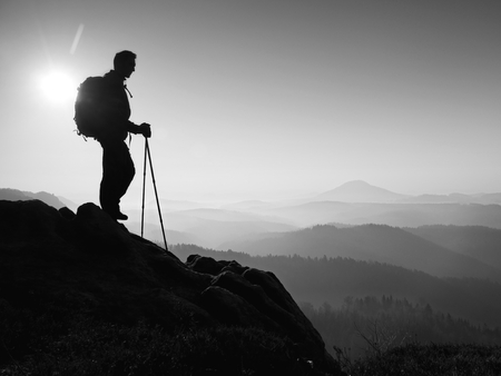 Tourist with sporty backpack and poles in hands stand on rocky view point and watching into deep misty valley bellow. Sunny spring daybreak in rocky mountains.の写真素材