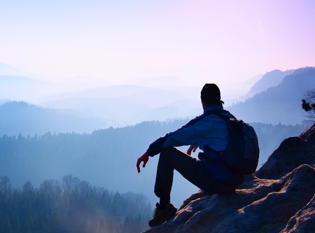 Tourist with sporty backpack sit on rocky peak  and watching into deep misty valley bellow. Sunny spring daybreak in rocky mountains.の写真素材