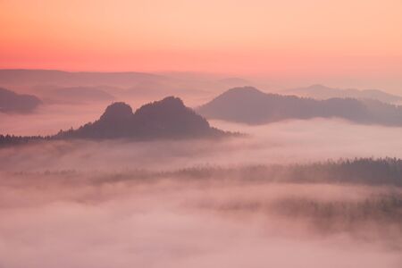 Red daybreak. Misty daybreak in a beautiful hills. Peaks of hills are sticking out from foggy background, the fog is red and orange due to Sun rays.の写真素材