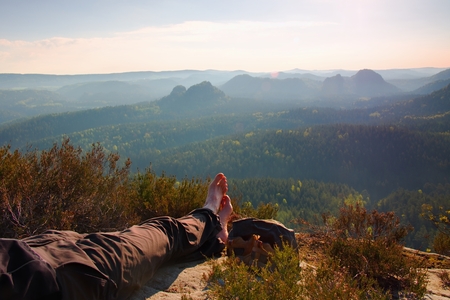 Long tired male legs in hiking trousers take a rest on peak of rock above valley.の写真素材