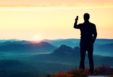 Man takes photos with smart phone on peak of rock empire. Dreamy fogy landscape, spring orange pink misty sunrise in a beautiful valley of rocky mountains.の写真素材