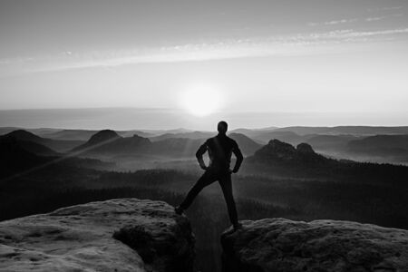 Hiker in black celebrate triumph between two rocky peaks. Wonderful daybreak, heavy mist in deep valleyの写真素材