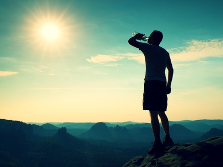 Thirsty hiker in black pants with bottle of water. Sweaty tired tourist on the peak of sandstone rocky park Saxony Switzerland watching into misty landscape.の写真素材