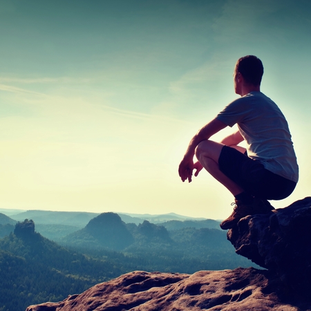 Young hiker in black pants and shirt  is sitting on cliff's edge and looking to misty hilly valley bellowの写真素材