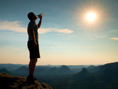 Thirsty hiker in black pants with bottle of water. Sweaty tired tourist on the peak of sandstone rocky park Saxony Switzerland watching into misty landscape.の写真素材