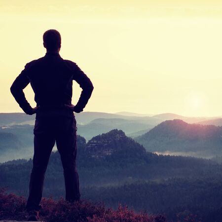 Gesture of triumph. Satisfy hiker in grey shirt and dark trousers. Tall man on the peak of cliff watching down to landscape.の写真素材