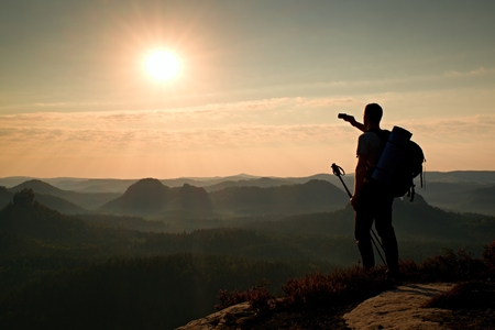 Tall backpacker with poles in hand. Sunny spring daybreak in rocky mountains. Hiker with big backpack stand on rocky view point above misty valley.の写真素材
