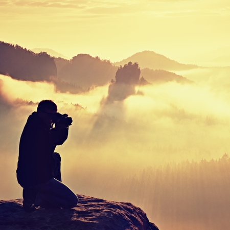 Happy photo enthusiast is  enjoying fantastic miracle of nature on cliff on rock. Dreamy fogy landscape, blue misty sunrise in a beautiful valley belowの写真素材