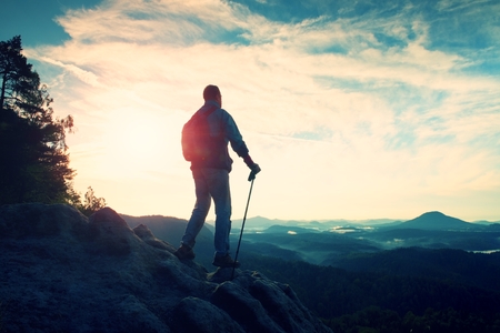 Tourist guide on the way with pole in hand. Hiker with sporty backpack stand on rocky view point above misty valley. Sunny spring daybreak in rocky mountains.の写真素材