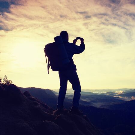 Tourist with backpack takes photos with smart phone on the rocky peak. Dreamy fogy valley belowの写真素材