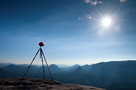 Tripod with red baseball cap on the peak ready for photography. Sharp autumn rocky peaks increased from gold foggy background,.の写真素材