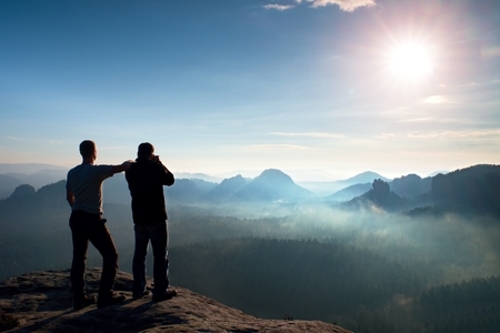 Hiker and photo enthusiast stay on cliff and thinking. Dreamy fogy landscape, blue misty sunrise in a beautiful valley belowの写真素材