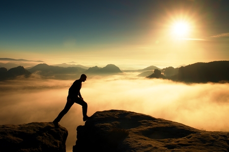 Hiker in black is jumping between the rocky peaks. Wonderful daybreak in rocky mountains, heavy orange mist in deep valley. Miracle of natureの写真素材