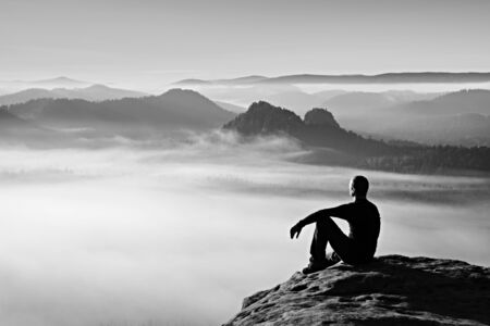 Hiker in black on the rocky peak. Wonderful daybreak in mountains, heavy mist in deep valley. Man sit on the rock.の写真素材