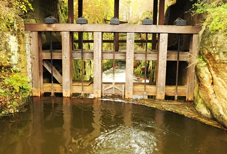 Water of small river flows at historical wooden weir. Terrible smell water with bubbles and reflection. Stony walls of Weirの写真素材